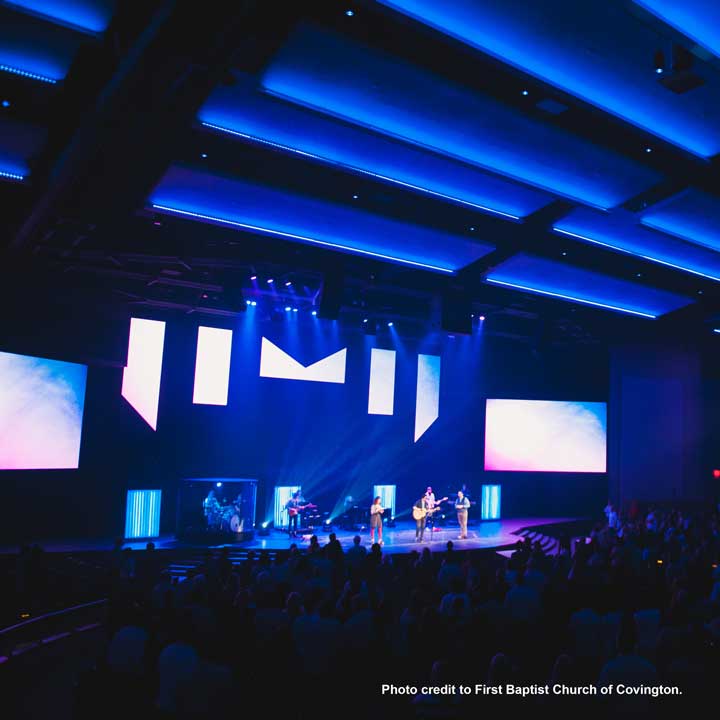A worship band performs on a brightly lit stage with dynamic lighting and large screens, while an audience watches in a dark auditorium and video captures the experience.