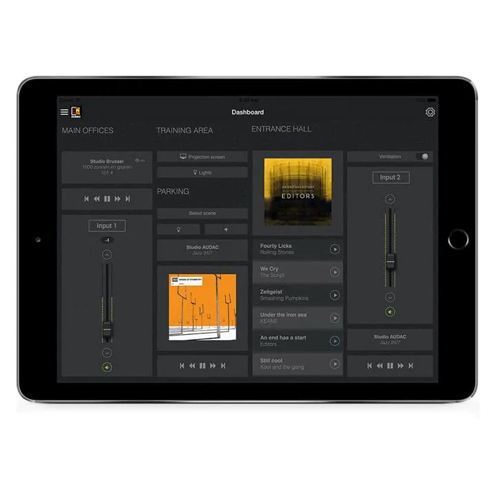 A black tablet displaying an audio control dashboard with labeled sections, audio track covers, volume sliders, and playback controls, alongside options for managing lighting on a dark background.