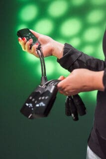 A person with red-painted nails holds multiple black audio and video devices with cables in both hands against a green background with circular light patterns.