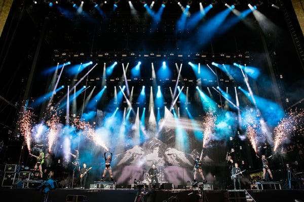 A rock band performs on a large stage with dramatic blue lighting and fireworks, set against a mountain backdrop, as various musicians play instruments under bright spotlight beams, creating a visually stunning video experience.