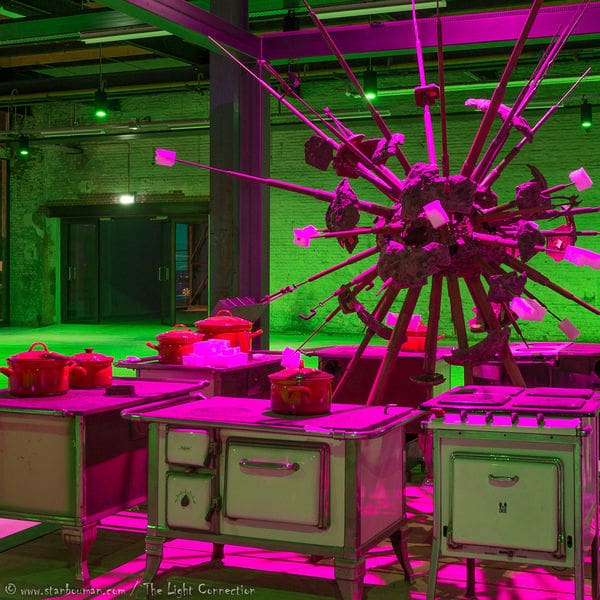 A room with vintage stoves is surrounded by a large spiked metal sculpture holding chunks of meat and fat. Dramatic green and pink lighting creates an unusual atmosphere, while red pots sit on the stoves, enhancing the visual scene like a vivid video.